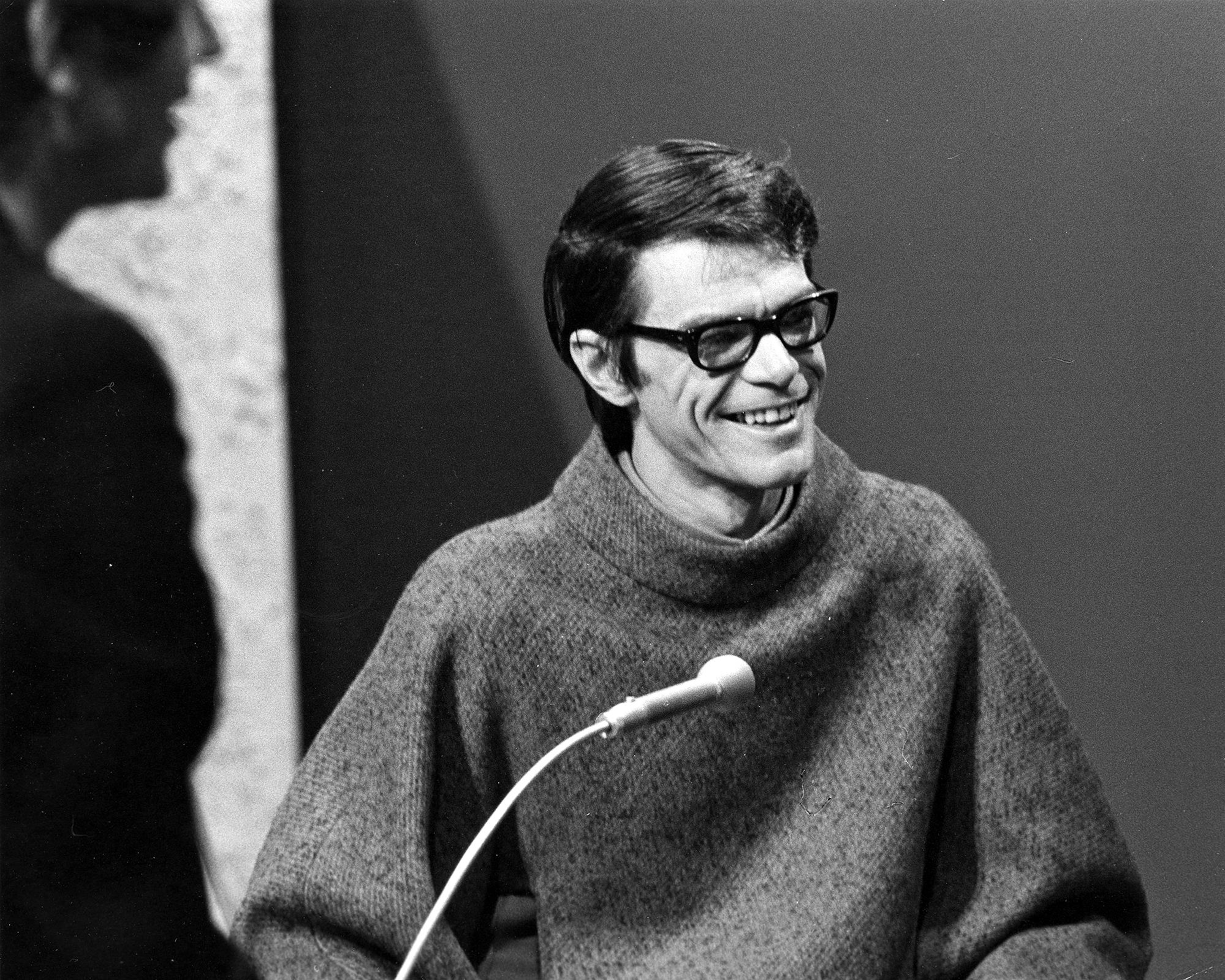 Photo en noir et blanc d'un homme portant des lunettes qui sourit devant un micro.