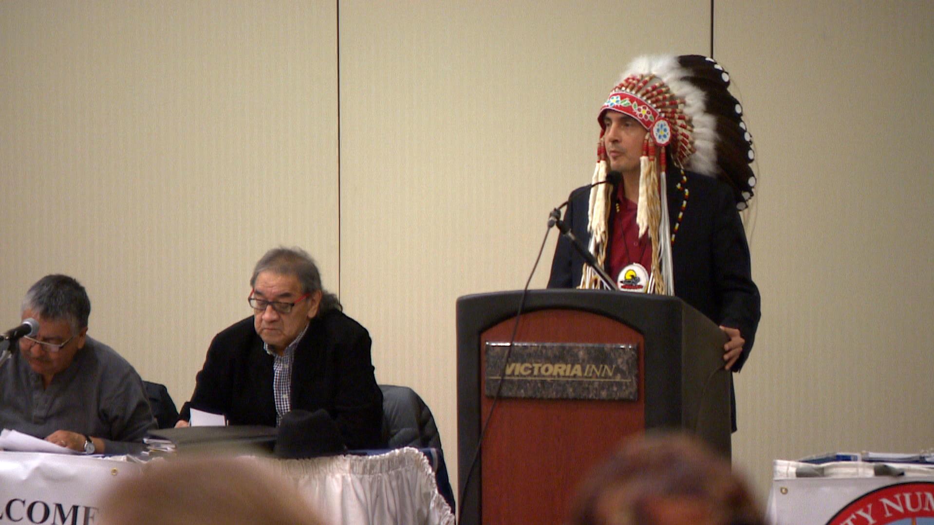 Un indígena canadiense hombre con un atuendo de plumas da un discurso desde un podio.