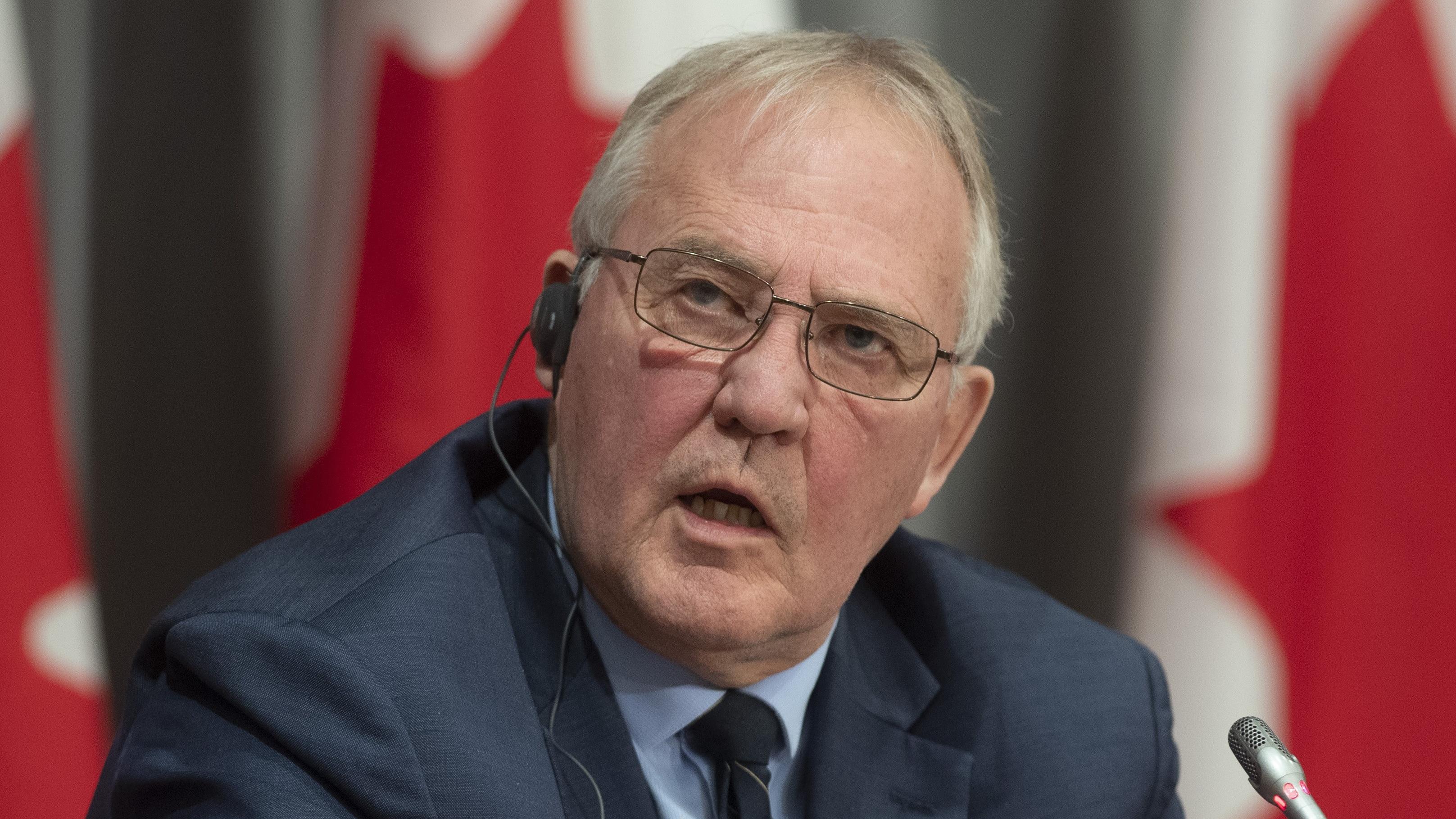 Bill Blair con un auricular en la oreja derecha, delante de banderas canadienses.