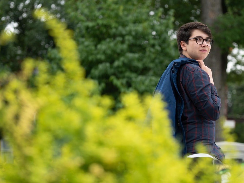 On voit Adam, debout, de profil, dans un parc. Il porte son manteau sur l'épaule.