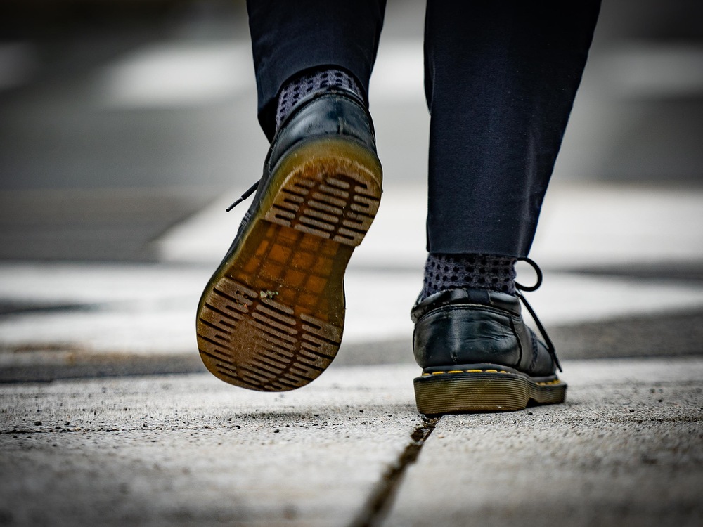 On voit en gros plan les pieds d'Adam, de derrière, qui marche sur le trottoir. Il porte des chaussures de cuir.