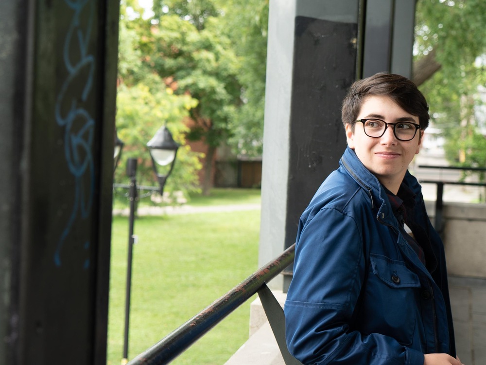 On voit Adam, debout, de profil, souriant. Il est adossé à une rambarde dans le kiosque d'un parc et porte un manteau.