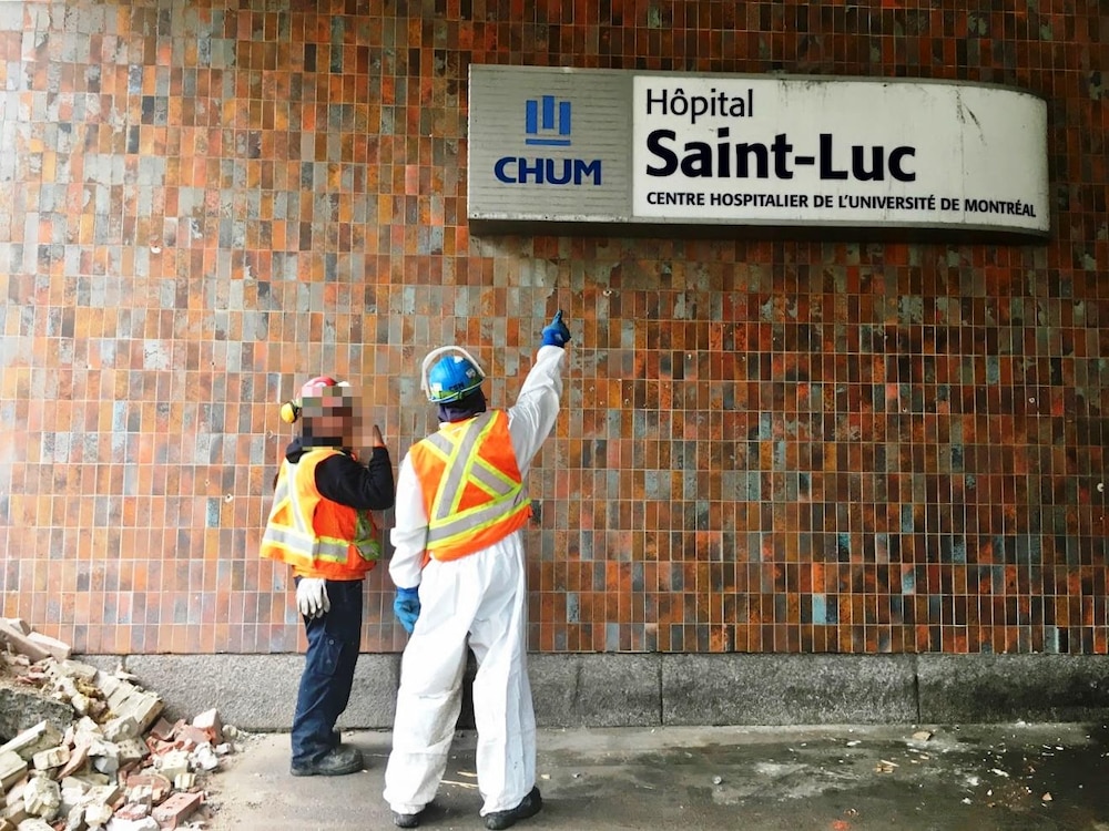 Des hommes, portant des casques de construction, regardent une affiche de l’hôpital Saint-Luc.