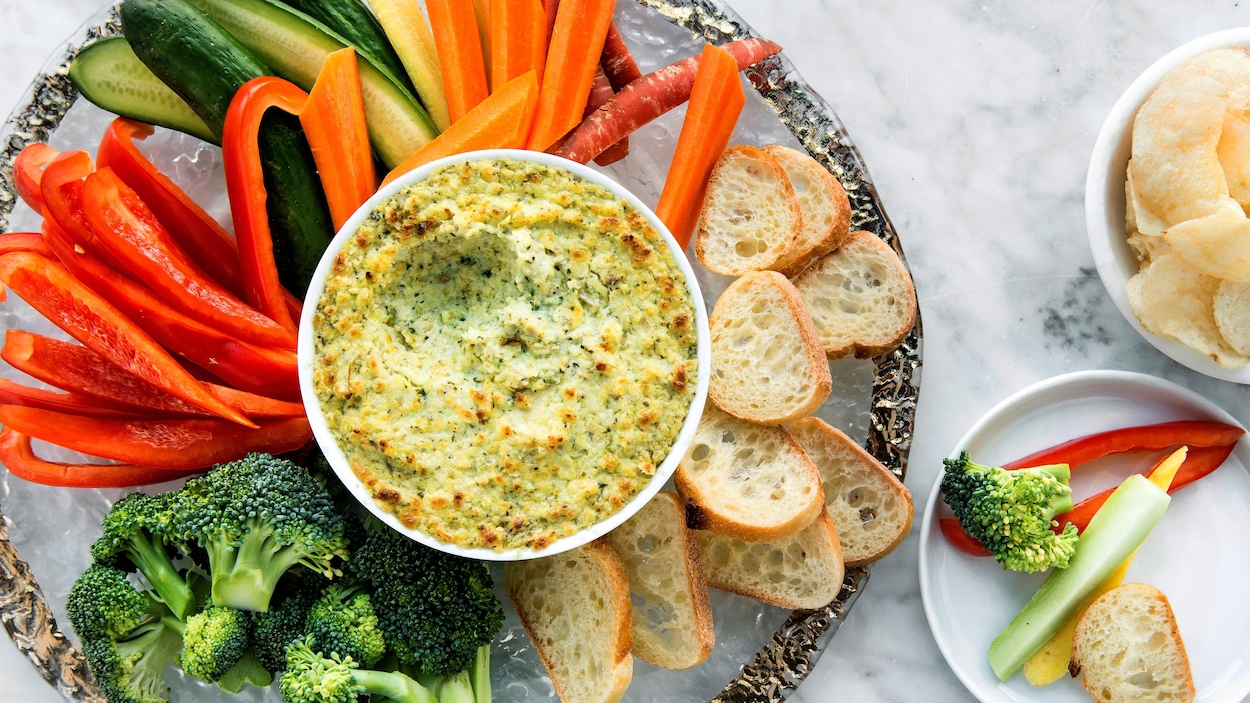 Trempette chaude aux artichauts dans un bol au milieu d'une assiette entouré de crudités et de tranches de baguette.