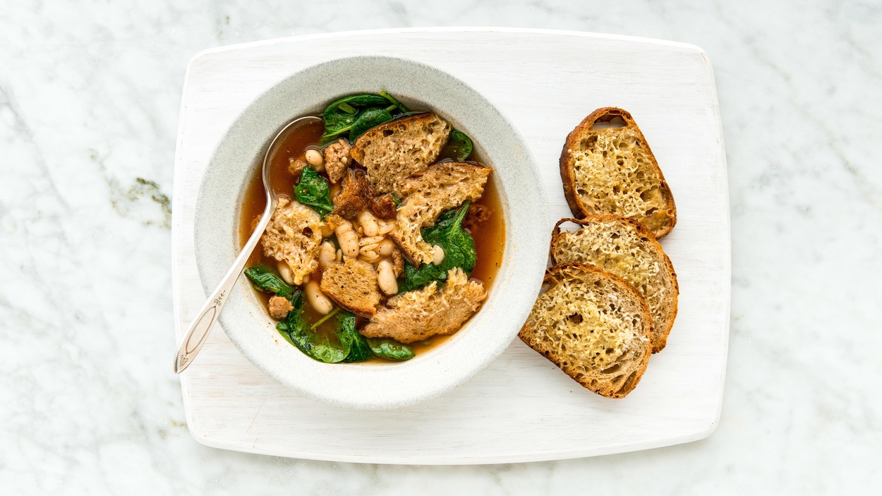 Un bol contenant une soupe aux saucisses et haricots blancs. Des croutons au parmesan sont disposés à la droite du bol de soupe.