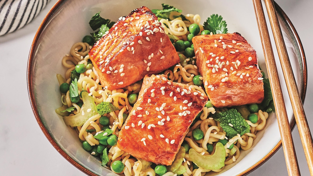 Des morceaux de saumon laqués à la sauce hoisin sont sur un nid de nouilles ramen garnies de petits pois et d'herbes fraîches.