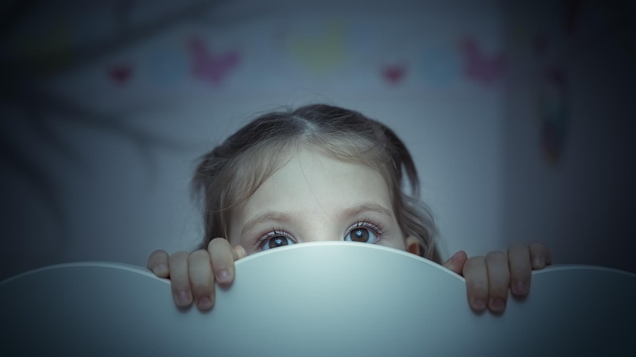 Une petite fille, à l'air effrayé, se cache derrière son lit. Une petite fille, à l'air effrayé, se cache derrière son lit.