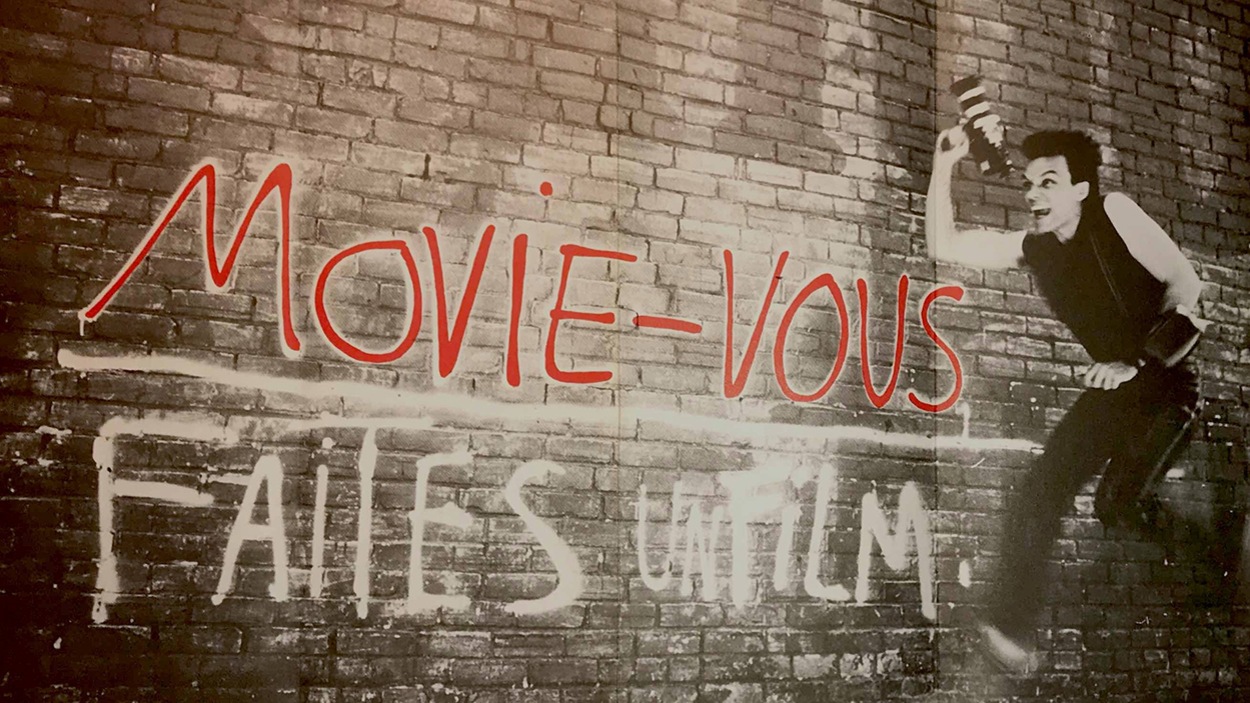 Un jeune homme tenant une caméra Super 8 se tient devant le slogan « Movie-vous, faites un film », peint sur un mur de briques. Un jeune homme tenant une caméra Super 8 se tient devant le slogan « Movie-vous, faites un film », peint sur un mur de briques.