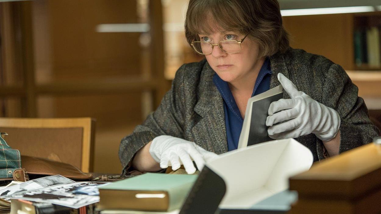 Une femme portant des lunettes et des gants, assise, manipule des livres devant elle. Une femme portant des lunettes et des gants, assise, manipule des livres devant elle.