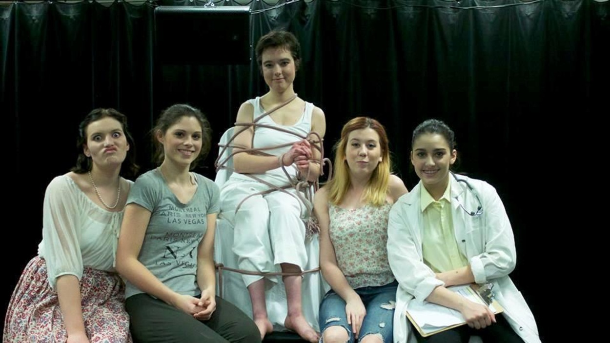 5 femmes sont sur scène. Celle du centre est attachée sur une chaise. 5 femmes sont sur scène. Celle du centre est attachée sur une chaise.