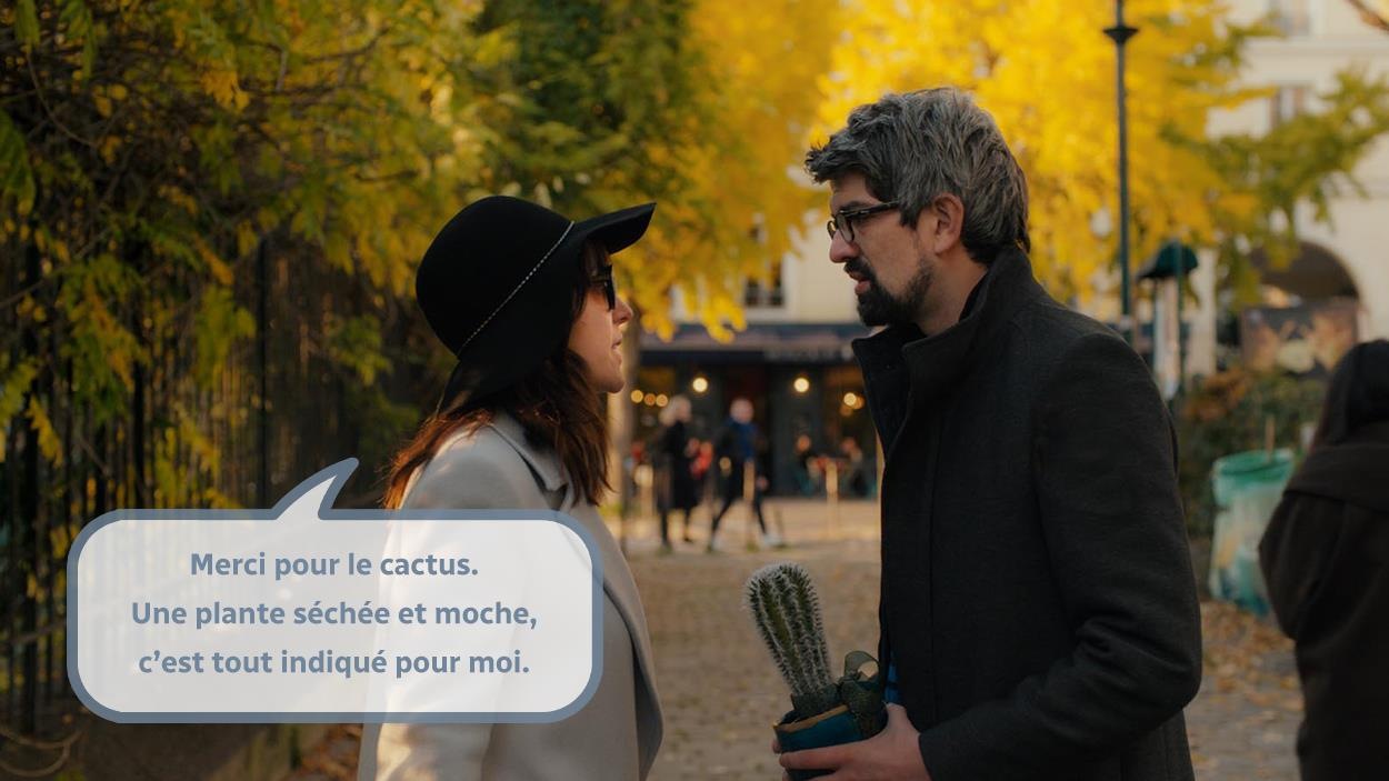 Une femme (Monia Chorki) portant un chapeau et des lunettes noires parle à un homme tenant un cactus. Une femme (Monia Chorki) portant un chapeau et des lunettes noires parle à un homme tenant un cactus.