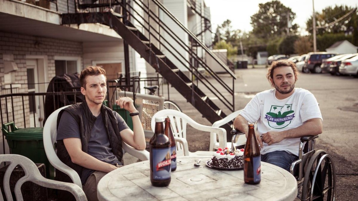 Deux jeunes hommes (dont Pier-Luc Funk et un autre en chaise roulante) assis autour d'une table, dehors, dans un parking. Deux jeunes hommes (dont Pier-Luc Funk et un autre en chaise roulante) assis autour d'une table, dehors, dans un parking.