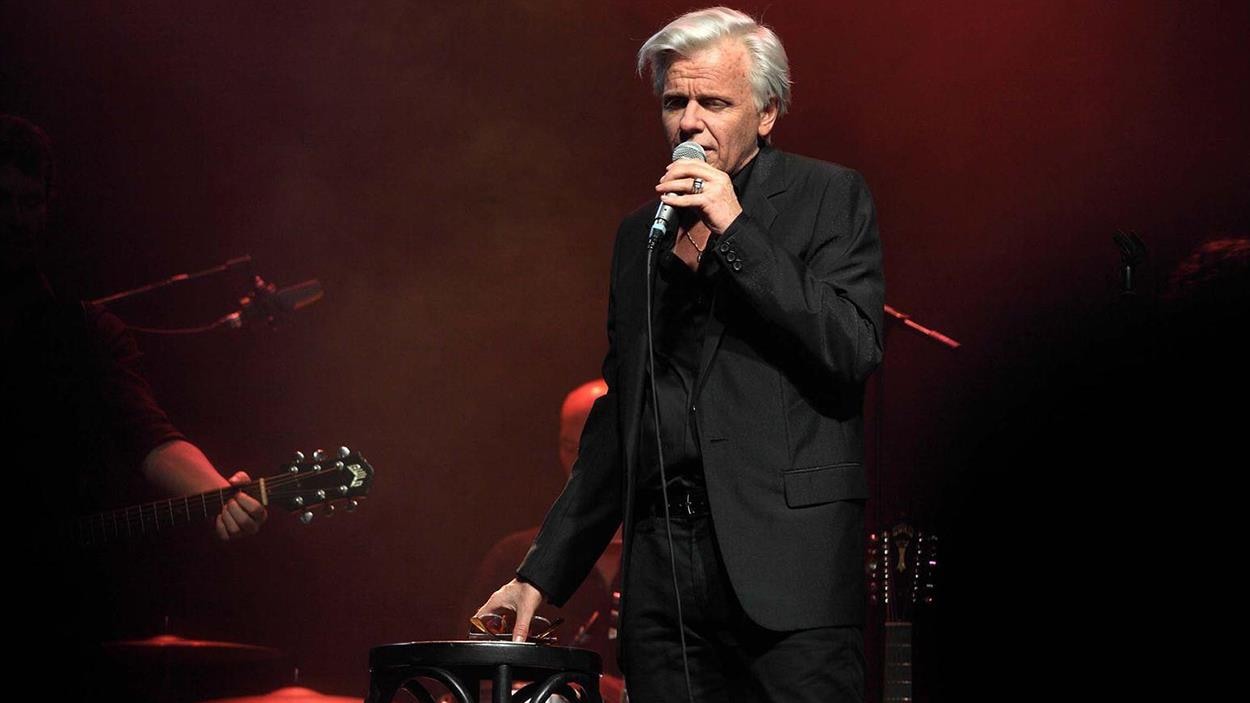 Un homme à la chevelure blanche, en costume sombre, sur scène en train de chanter. Un homme à la chevelure blanche, en costume sombre, sur scène en train de chanter.