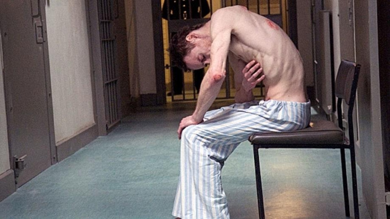 Un homme décharné, le torse nu, en pantalon rayé, assis sur une chaise dans un couloir de prison Un homme décharné, le torse nu, en pantalon rayé, assis sur une chaise dans un couloir de prison