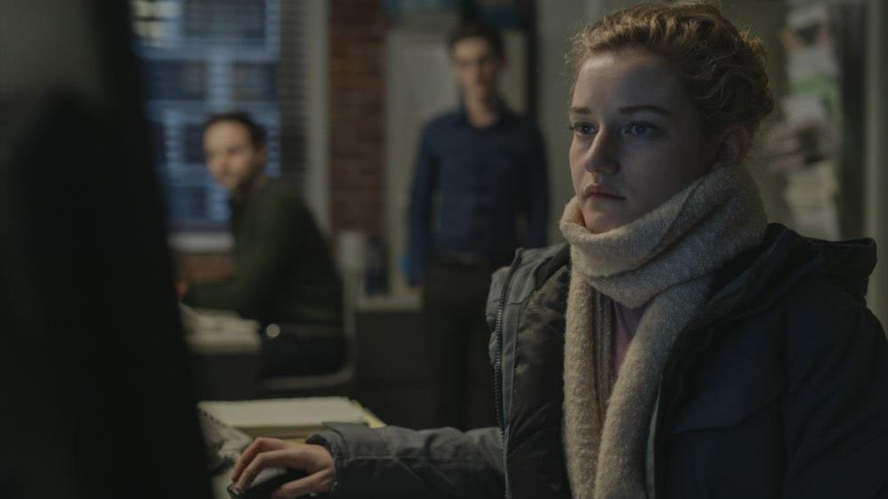 Une jeune fille portant manteau et écharpe tape à son ordinateur, devant deux de ses collègues masculins en arrière-plan.