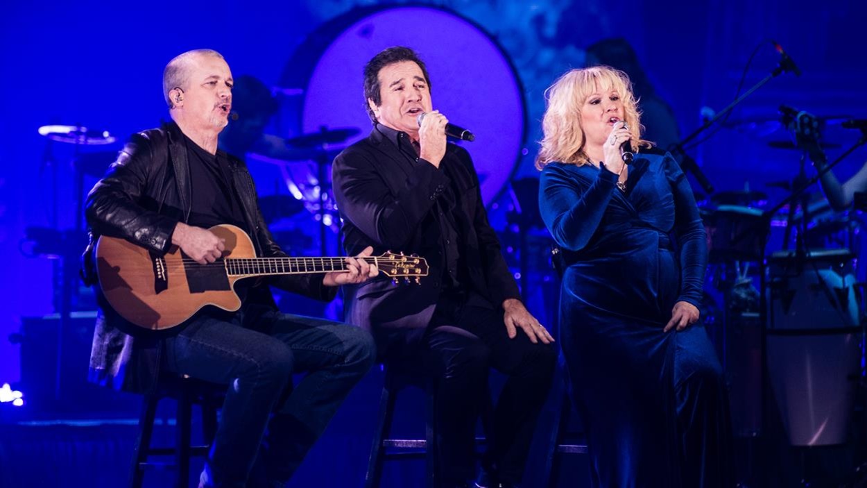 Les trois chanteurs sont assis sur scène, une guitare ou micro à la main. Les trois chanteurs sont assis sur scène, une guitare ou micro à la main.