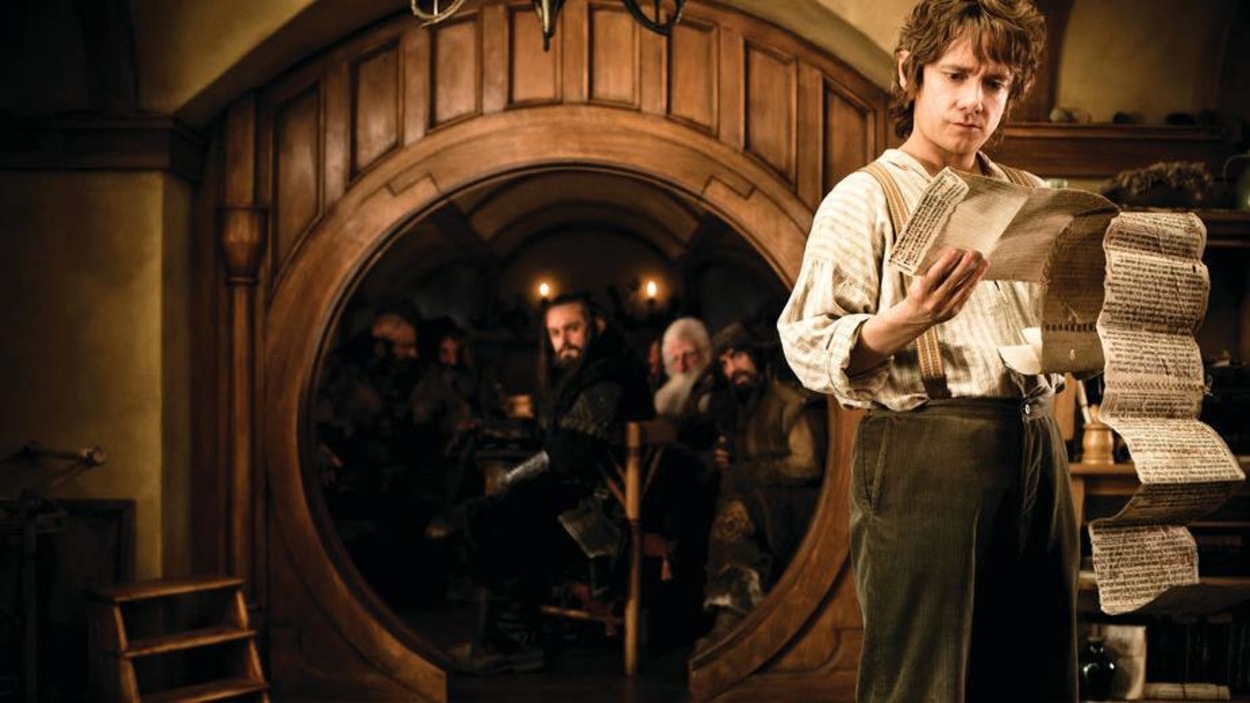 Un hobbit lit une longue lettre devant un cadre de porte rond sous lequel plusieurs hommes sont assis. Un hobbit lit une longue lettre devant un cadre de porte rond sous lequel plusieurs hommes sont assis.