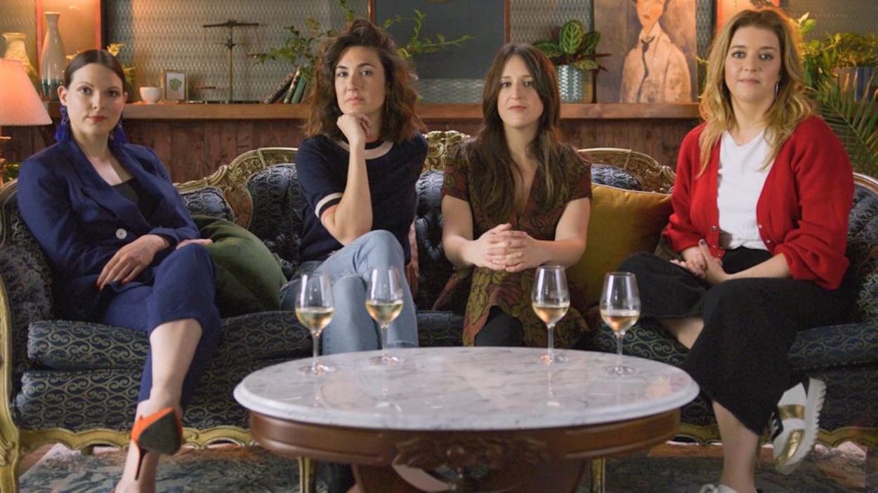Les 4 filles sont assises sur un élégant canapé. Des verres de vin blanc sont posés sur une table basse devant elles.