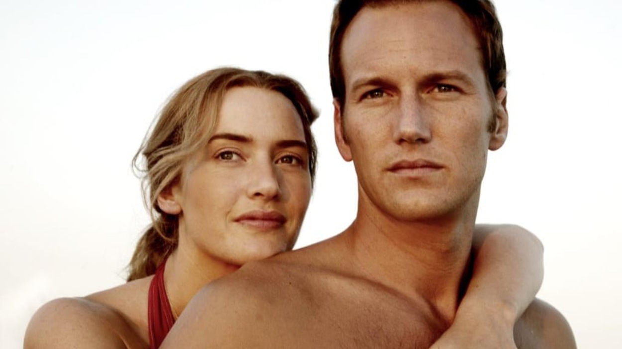 Une femme (Kate Winslet) en maillot rouge enlace par derrière un homme (Patrick Wilson) torse nu. Une femme (Kate Winslet) en maillot rouge enlace par derrière un homme (Patrick Wilson) torse nu.