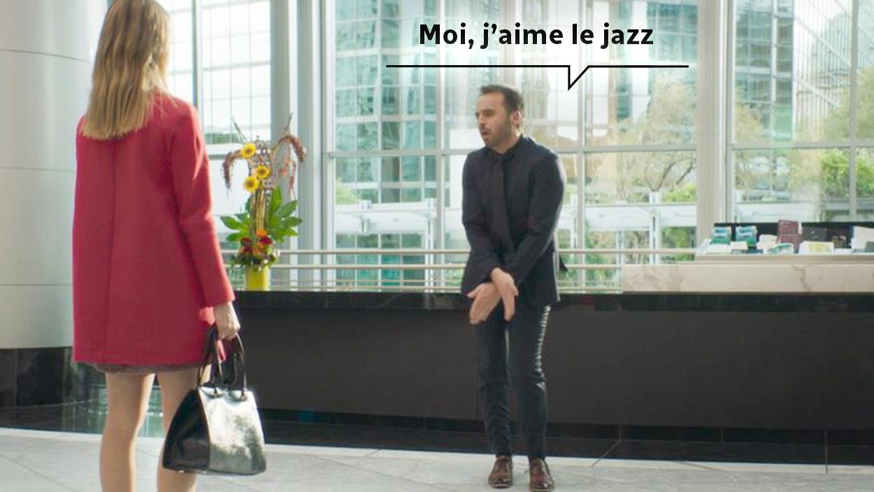 Face à une femme de dos, un homme (Louis-José Houde) fait un mouvement de danse et un dialogue écrit qui dit: Moi, j'aime le jazz.