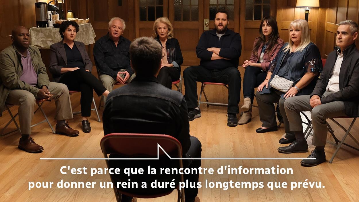 Un homme (Louis-José Houde) de dos face à un groupe de personnes assises et le dialogue écrit: C'est parce que la rencontre d'information pour donner un rein a duré plus longtemps que prévu.