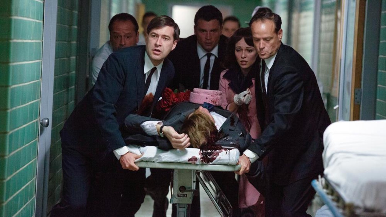 Un groupe d'hommes et une femme en tailleur rose autour d'une civière portant un corps ensanglanté dans un couloir d'hôpital. Un groupe d'hommes et une femme en tailleur rose autour d'une civière portant un corps ensanglanté dans un couloir d'hôpital.