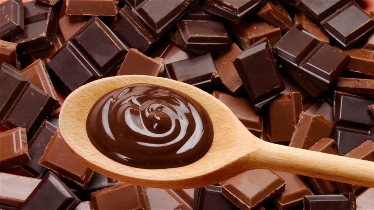 Un gros plan sur une cuillère de bois contenant du chocolat fondu. En arrière-plan, des carrés de chocolat noir et de chocolat au lait.