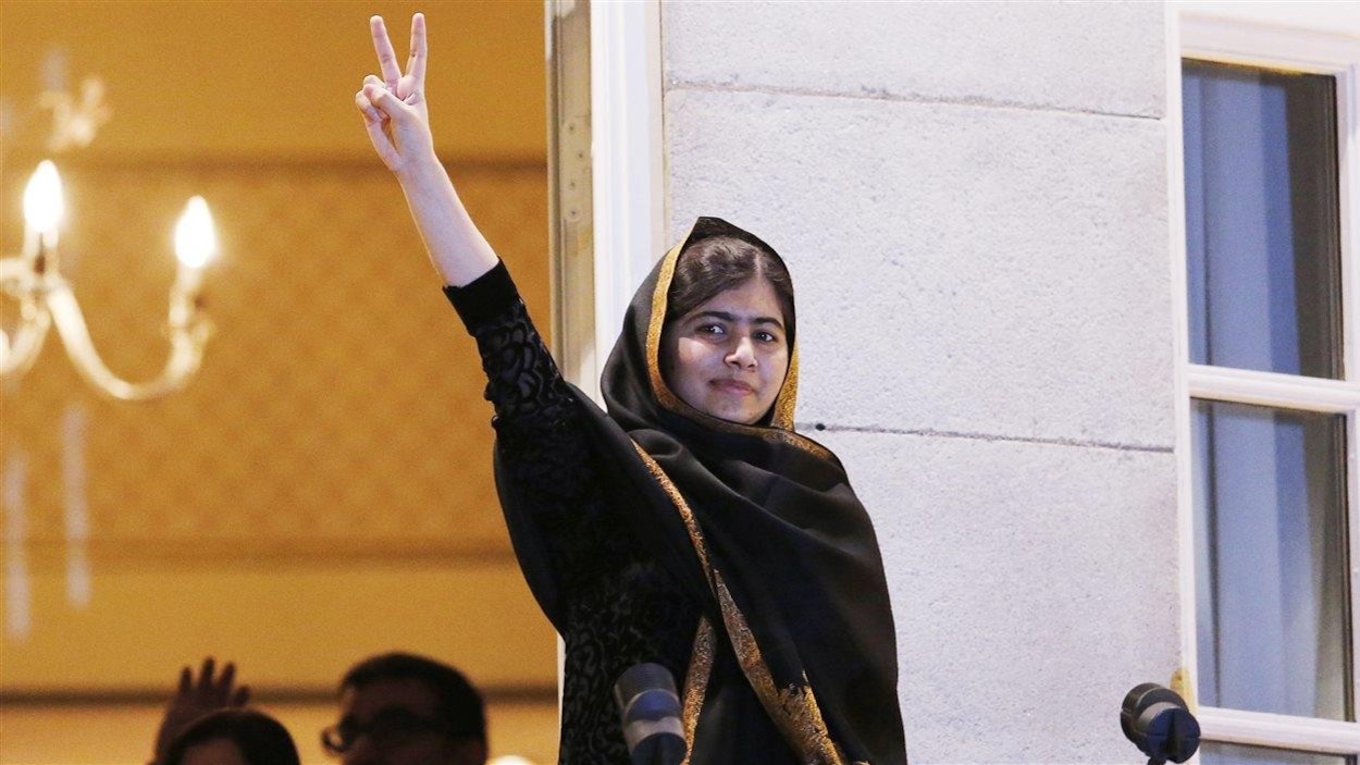 Malala Yousafzai, avec un léger sourire, est au balcon du Grand Hotel d'Oslo. Elle salue la foule en levant le bras et faisant le signe de la paix
