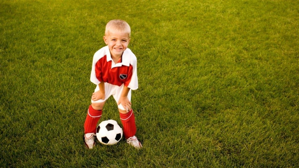 Un jeune garçon, en habit de soccer, est sur le terrain gazonné et regarde la caméra. Il a un ballon de soccer à ses pieds.