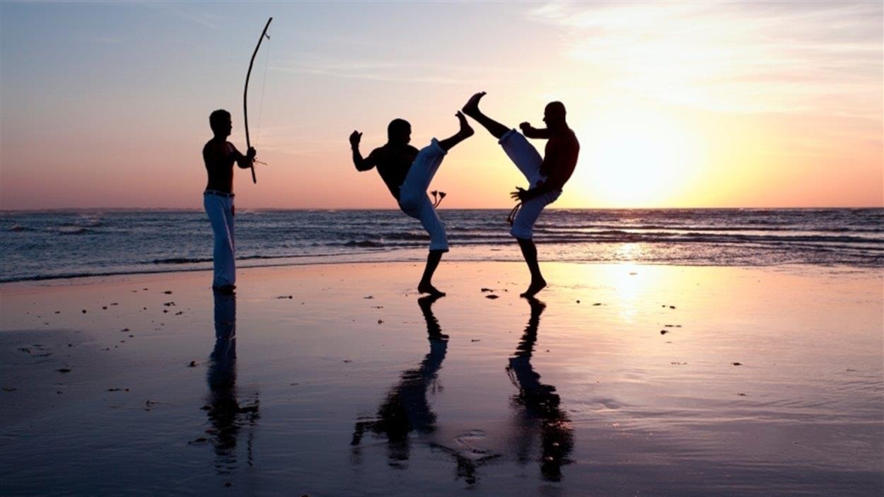 Deux hommes se battent en se donnant des coups de pied sur une plage. Un autre se tient debout et joue du bérimbau. 