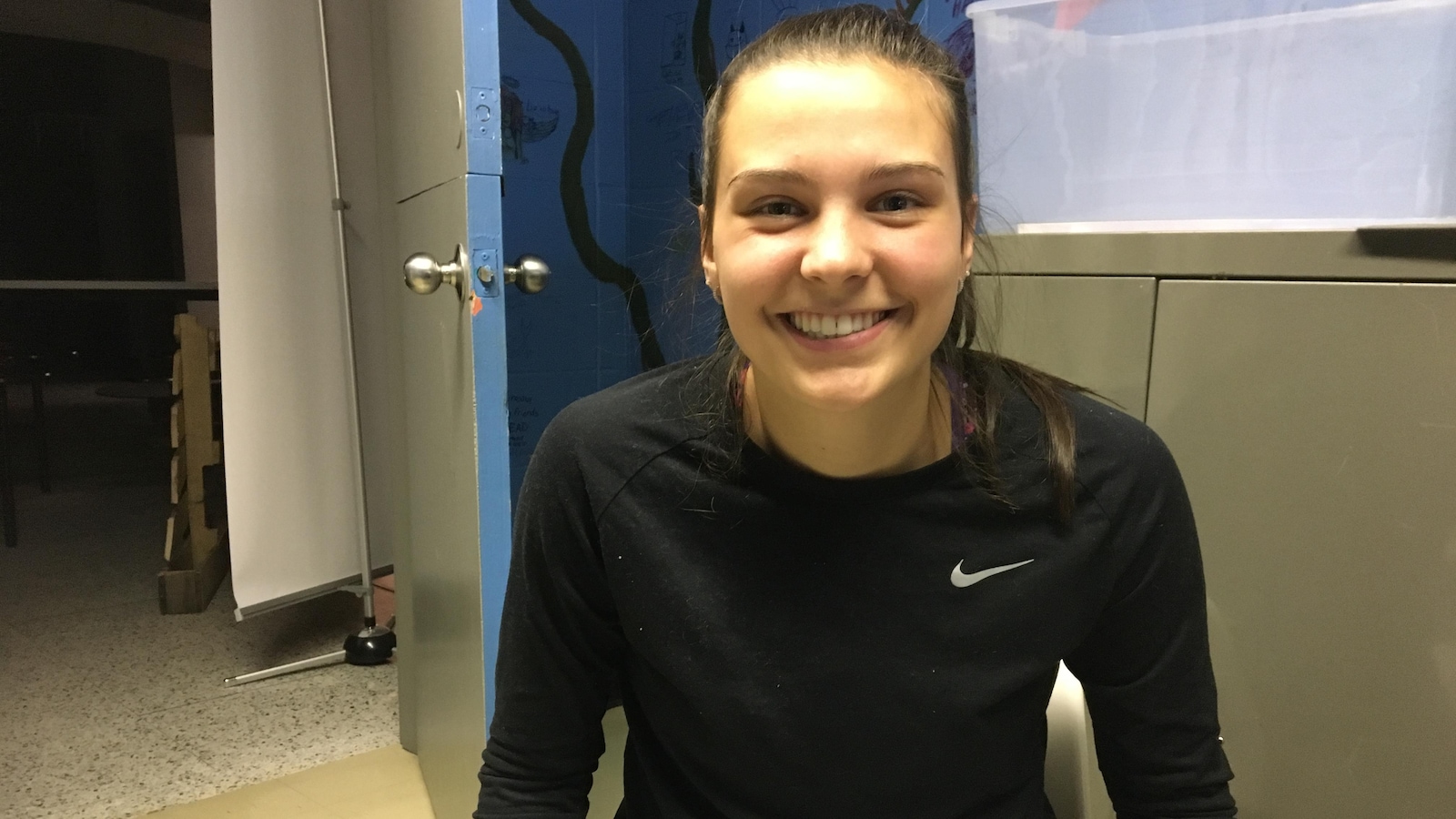 Une adolescente sourit à la caméra, assise dans un local de son école secondaire. Une adolescente sourit à la caméra, assise dans un local de son école secondaire.