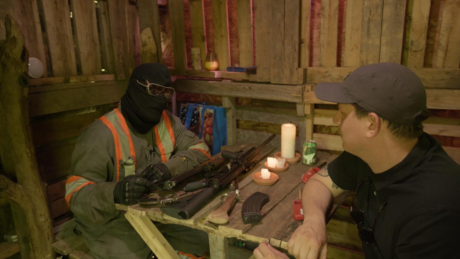 Deux hommes échangent autour d'une table couverte de fusils, dans une cabane en bois. 
