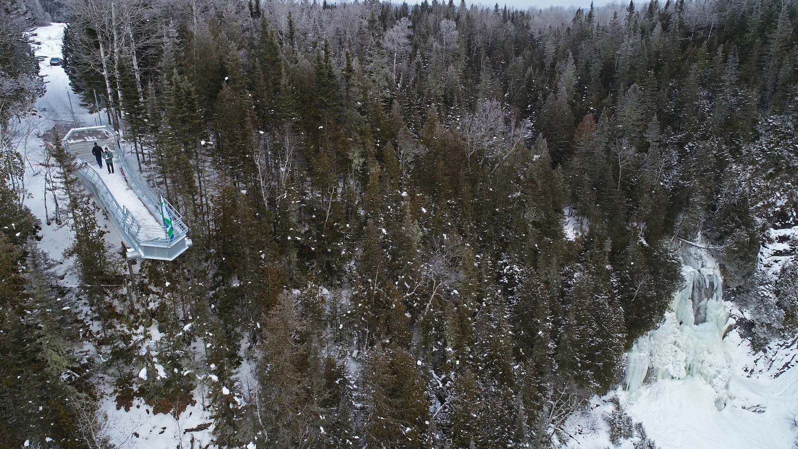 On voit un belvédère qui surplombe une falaise en plein hiver. Des conifères entourent la structure. Deux personnes sont sur la structure. On voit un belvédère qui surplombe une falaise en plein hiver. Des conifères entourent la structure. Deux personnes sont sur la structure.