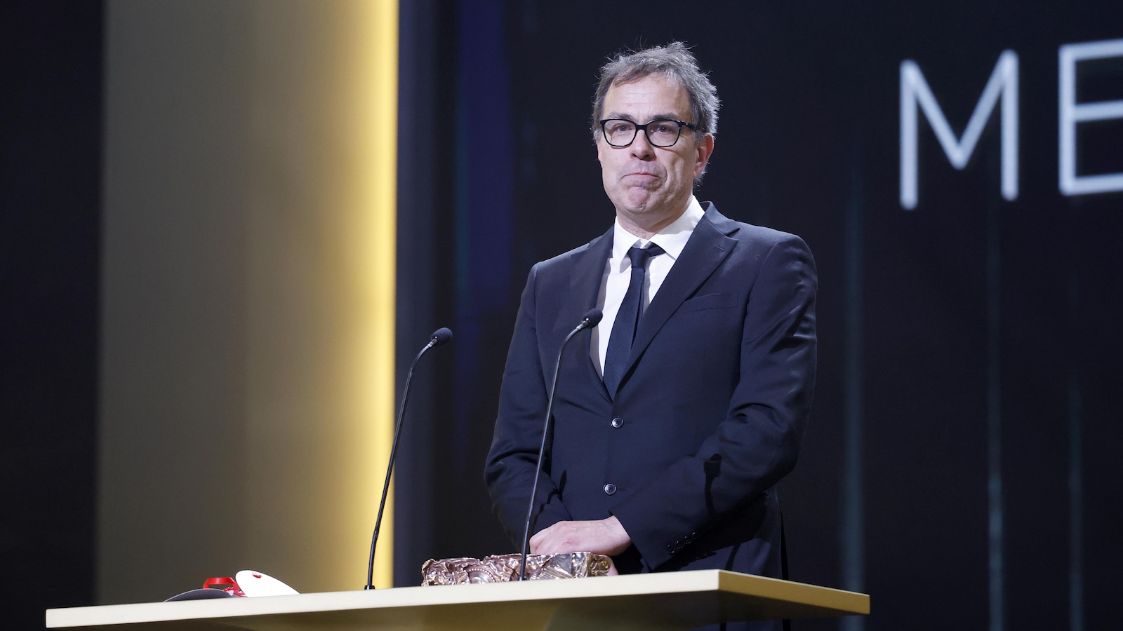 Un homme se tient debout sur une scène, face à un lutrin sur lequel est posé un trophée. Un homme se tient debout sur une scène, face à un lutrin sur lequel est posé un trophée.
