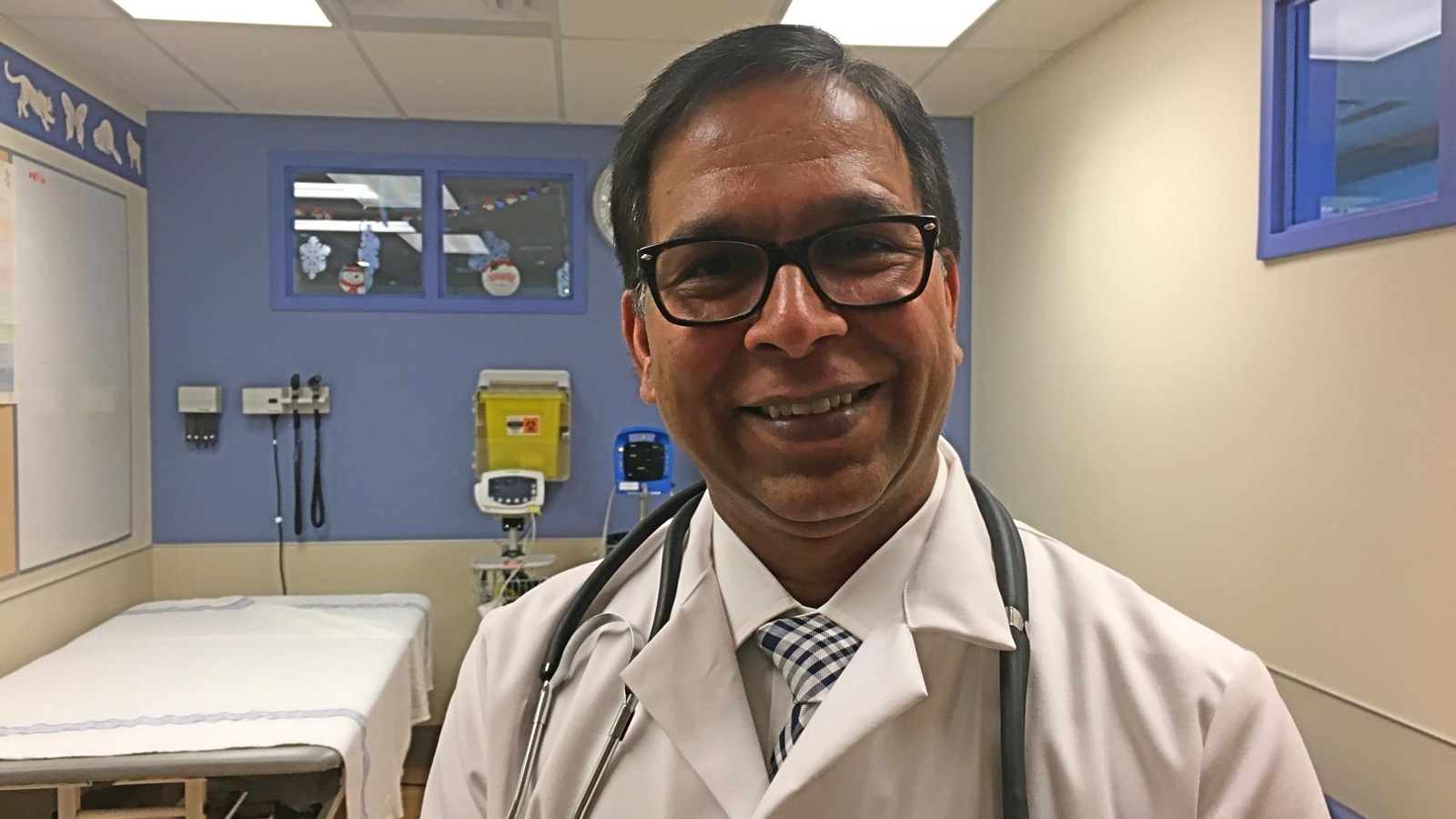 Un docteur en blouse blanche porte un stéthoscope autour du cou. Il pose, tout sourire, dans une salle d'examen d'un hôpital. Un docteur en blouse blanche porte un stéthoscope autour du cou. Il pose, tout sourire, dans une salle d'examen d'un hôpital.