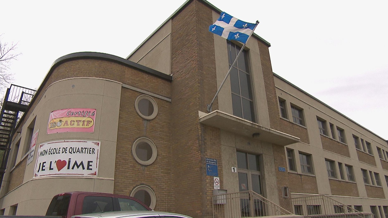 Un bâtiment en brique compte trois étages. Il y a une affiche sur la façade qui dit: Mon école de quartier je l'aime. Un bâtiment en brique compte trois étages. Il y a une affiche sur la façade qui dit: Mon école de quartier je l'aime.