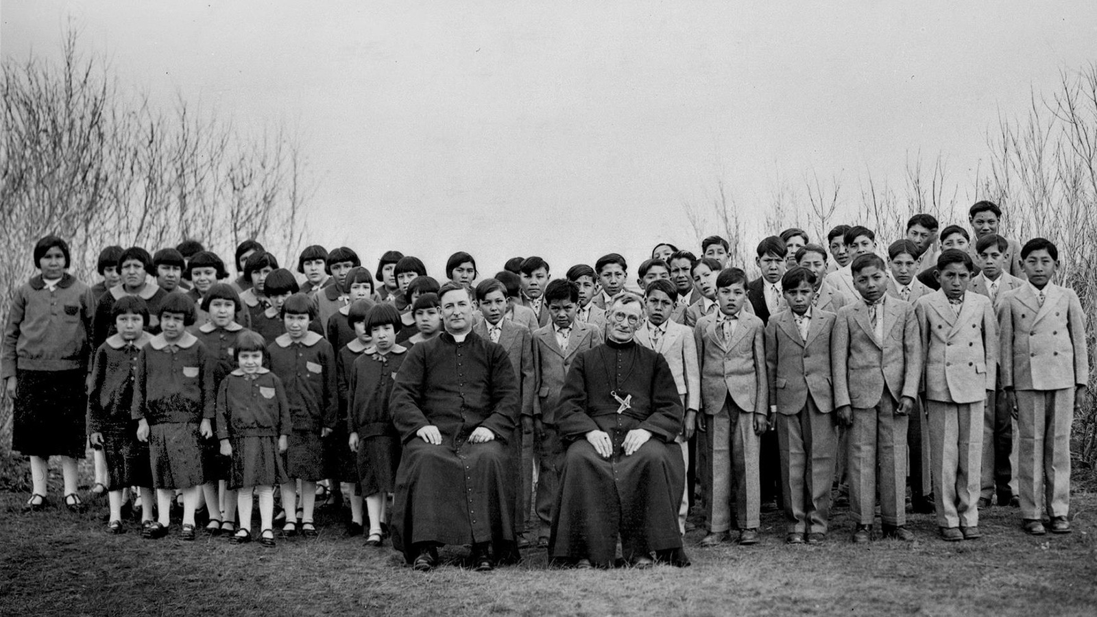 Des élèves du pensionnat autochtone Brocket, en Alberta, en 1930, sont debout en uniformes d'élèves derrière deux religieux assis. Des élèves du pensionnat autochtone Brocket, en Alberta, en 1930, sont debout en uniformes d'élèves derrière deux religieux assis.
