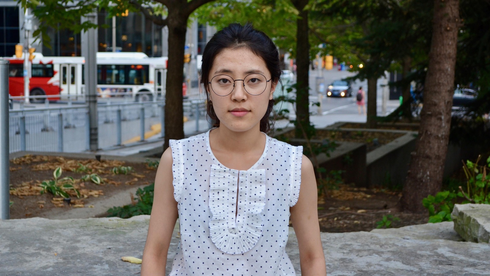 Une jeune femme portant des lunettes photographiée dans le centre-ville d'Ottawa, une rue et un autobus derrière elle Une jeune femme portant des lunettes photographiée dans le centre-ville d'Ottawa, une rue et un autobus derrière elle