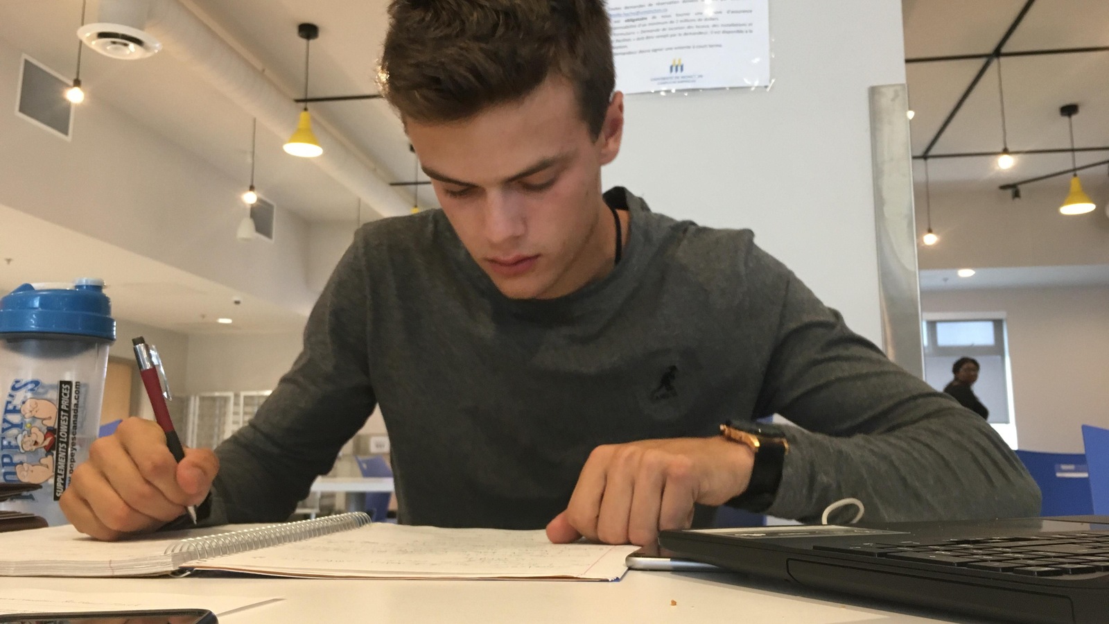 Un jeune attablé rédige des notes non loin de son ordinateur portable. Un jeune attablé rédige des notes non loin de son ordinateur portable.