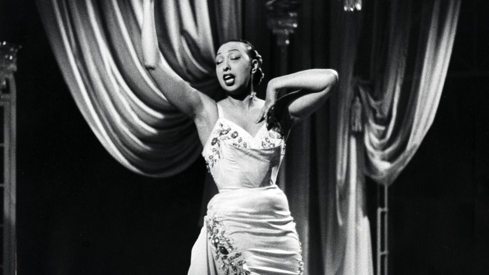Joséphine Baker chante et gesticule dans un décor de scène. Joséphine Baker chante et gesticule dans un décor de scène.