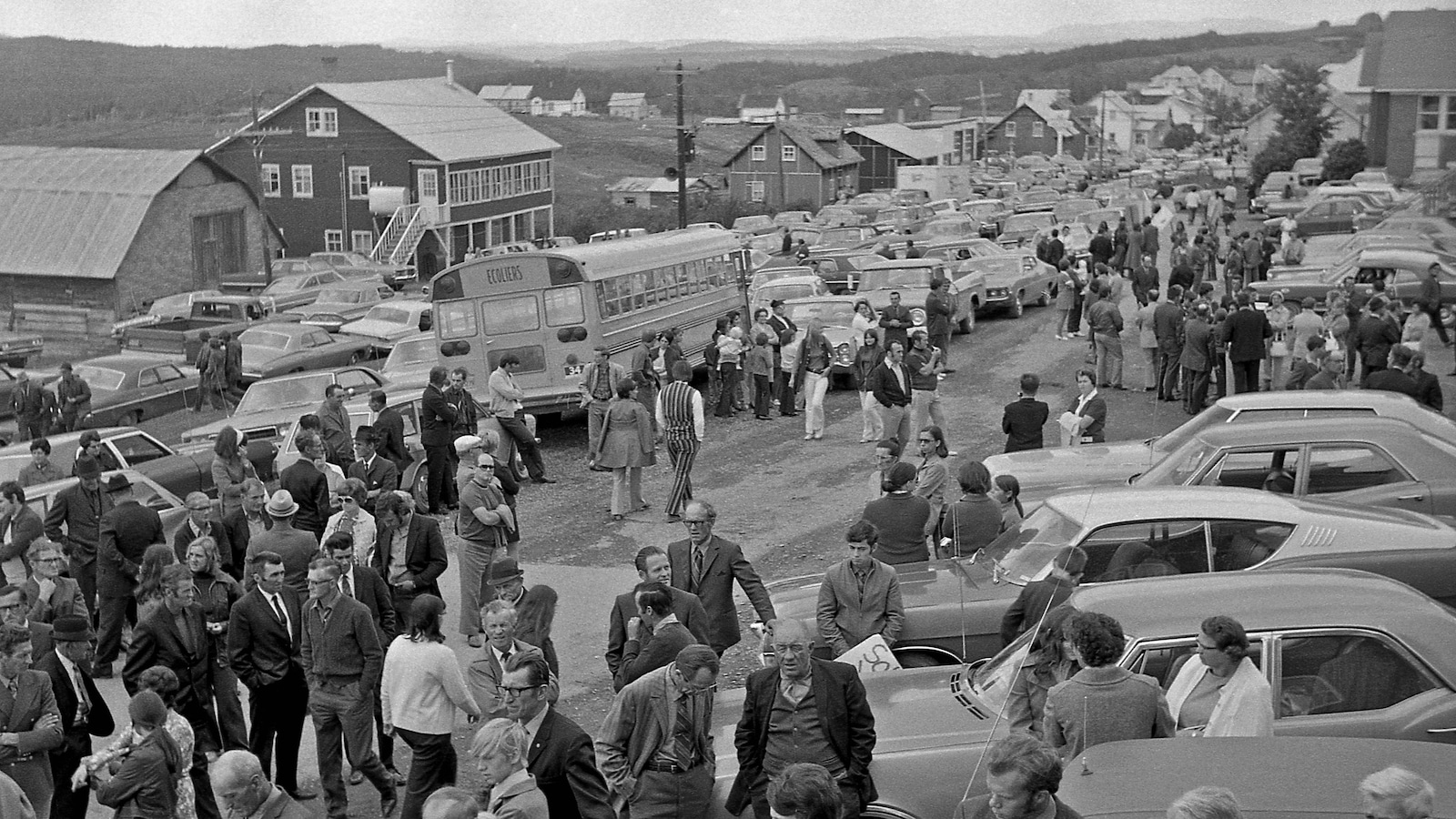 Une photo en noir et blanc de personnes qui sont dans une rue d'un village et se rassemblent. Des voitures sont stationnées à droite. Une photo en noir et blanc de personnes qui sont dans une rue d'un village et se rassemblent. Des voitures sont stationnées à droite.