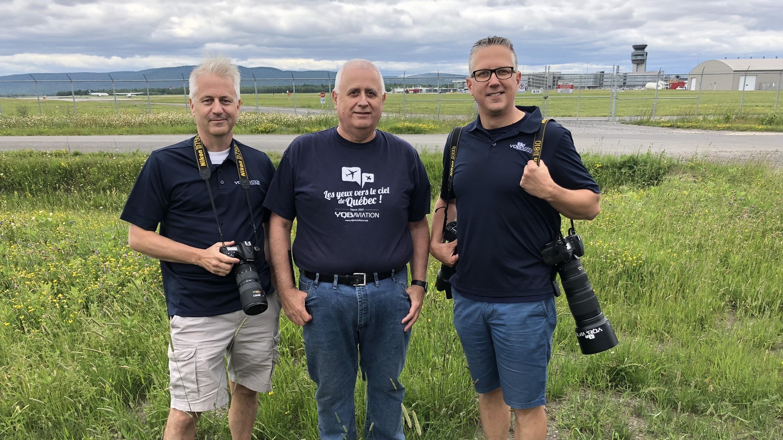 Trois hommes munis d'appareils photo se tiennent dans un champ devant l'aéroport Jean-Lesage de Québec. Trois hommes munis d'appareils photo se tiennent dans un champ devant l'aéroport Jean-Lesage de Québec.
