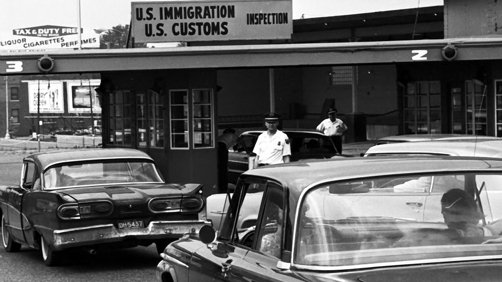 Photo en noir et blanc. Des voitures sont arrêtées à un poste frontière. Des agents sont dehors Photo en noir et blanc. Des voitures sont arrêtées à un poste frontière. Des agents sont dehors