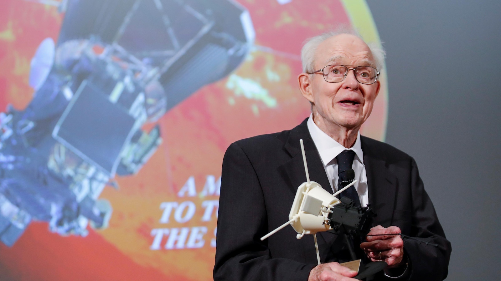 Un scientifique tenant une maquette devant une projection d'un logo de mission spatiale. Un scientifique tenant une maquette devant une projection d'un logo de mission spatiale.