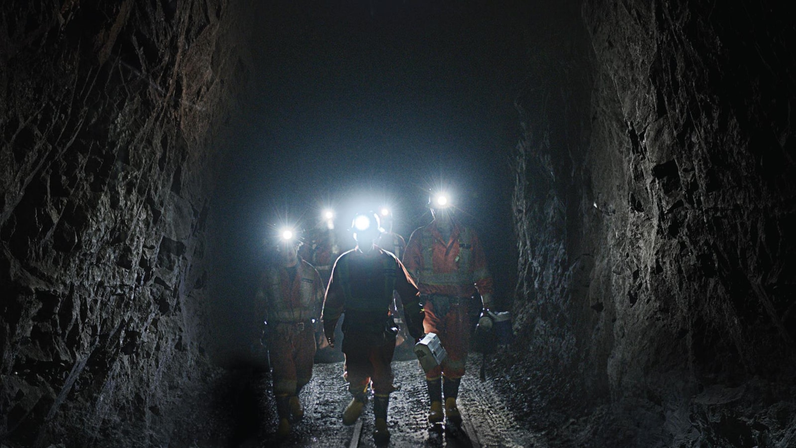 Cinq mineurs et mineures se promènent dans une mine, portant des casques équippés d'une lampe torche. 