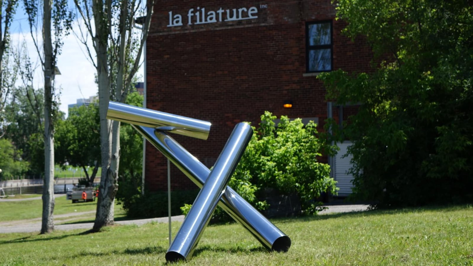 La photo illustre une oeuvre d'art composée de trois tubes en acier inoxydable, exposée devant un édifice historique. La photo illustre une oeuvre d'art composée de trois tubes en acier inoxydable, exposée devant un édifice historique.