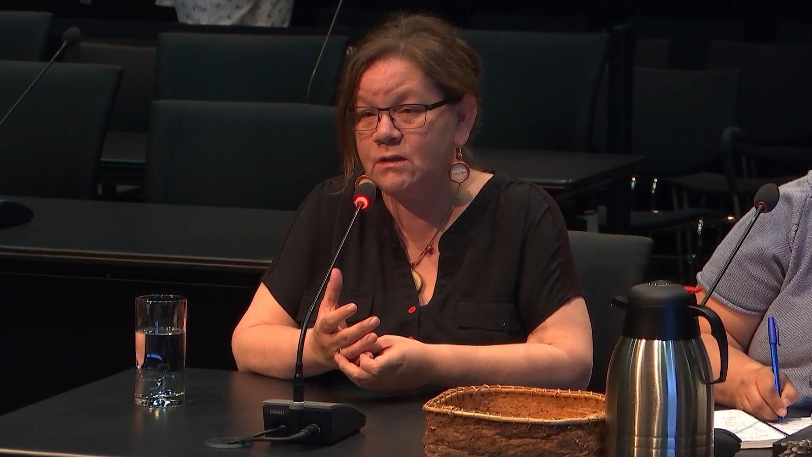 Une femme aux cheveux bruns qui sont attachés porte des minces lunettes noires et parle dans un micro noir posé sur le bureau devant elle. Une femme aux cheveux bruns qui sont attachés porte des minces lunettes noires et parle dans un micro noir posé sur le bureau devant elle.