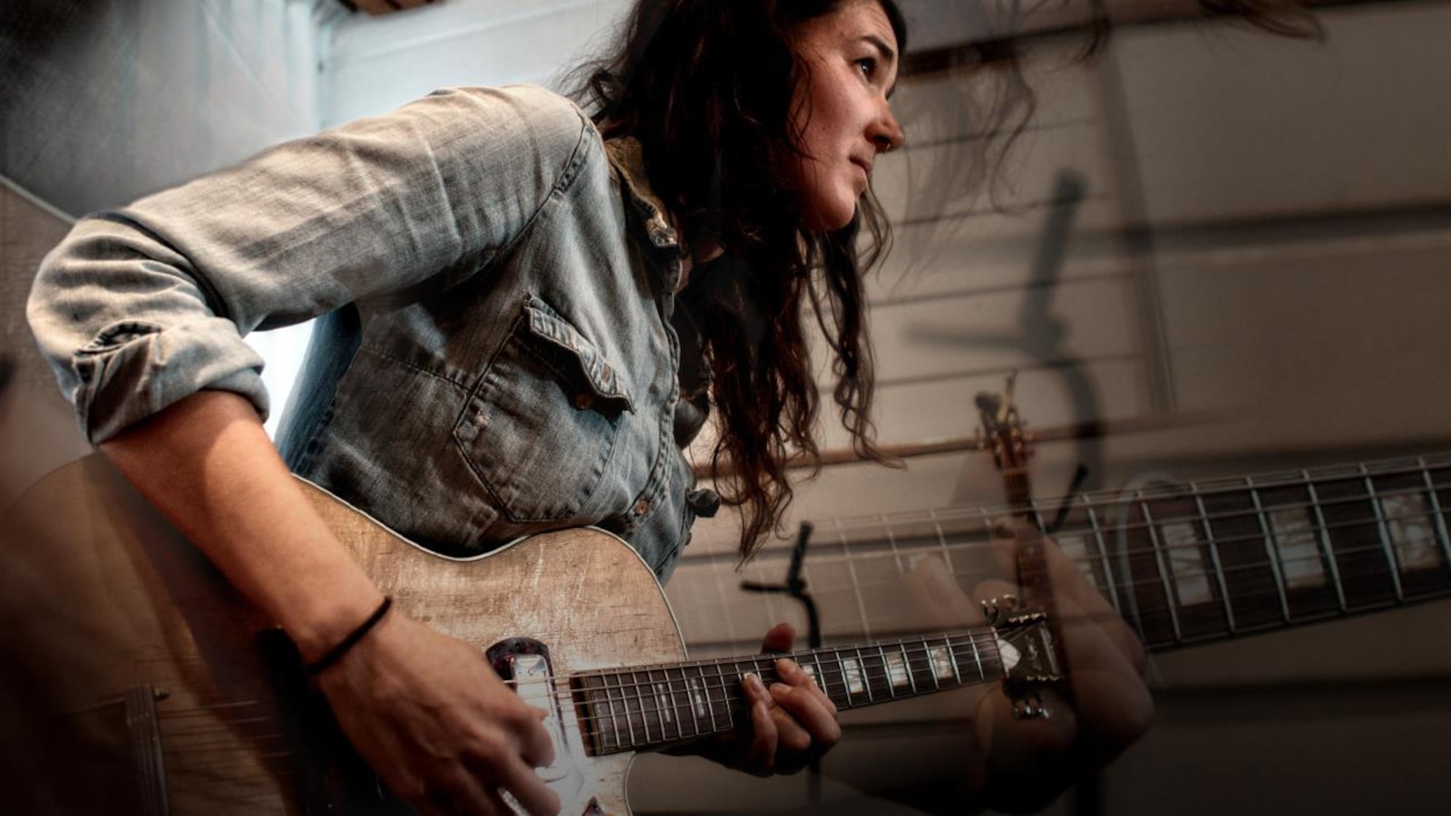 Maggie Savoie à la guitare dans ce qui semble être un studio. Elle est de profile et porte une chemise en jeans. Maggie Savoie à la guitare dans ce qui semble être un studio. Elle est de profile et porte une chemise en jeans.