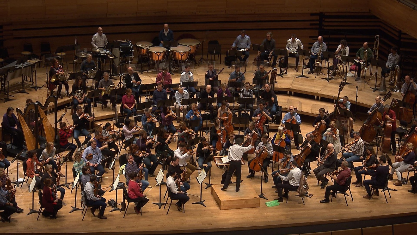 On voit, en plongée depuis un balcon, Kent Nagano et les membres de l'OSM en pleine répétition à la Maison symphonique de Montréal. On voit, en plongée depuis un balcon, Kent Nagano et les membres de l'OSM en pleine répétition à la Maison symphonique de Montréal.