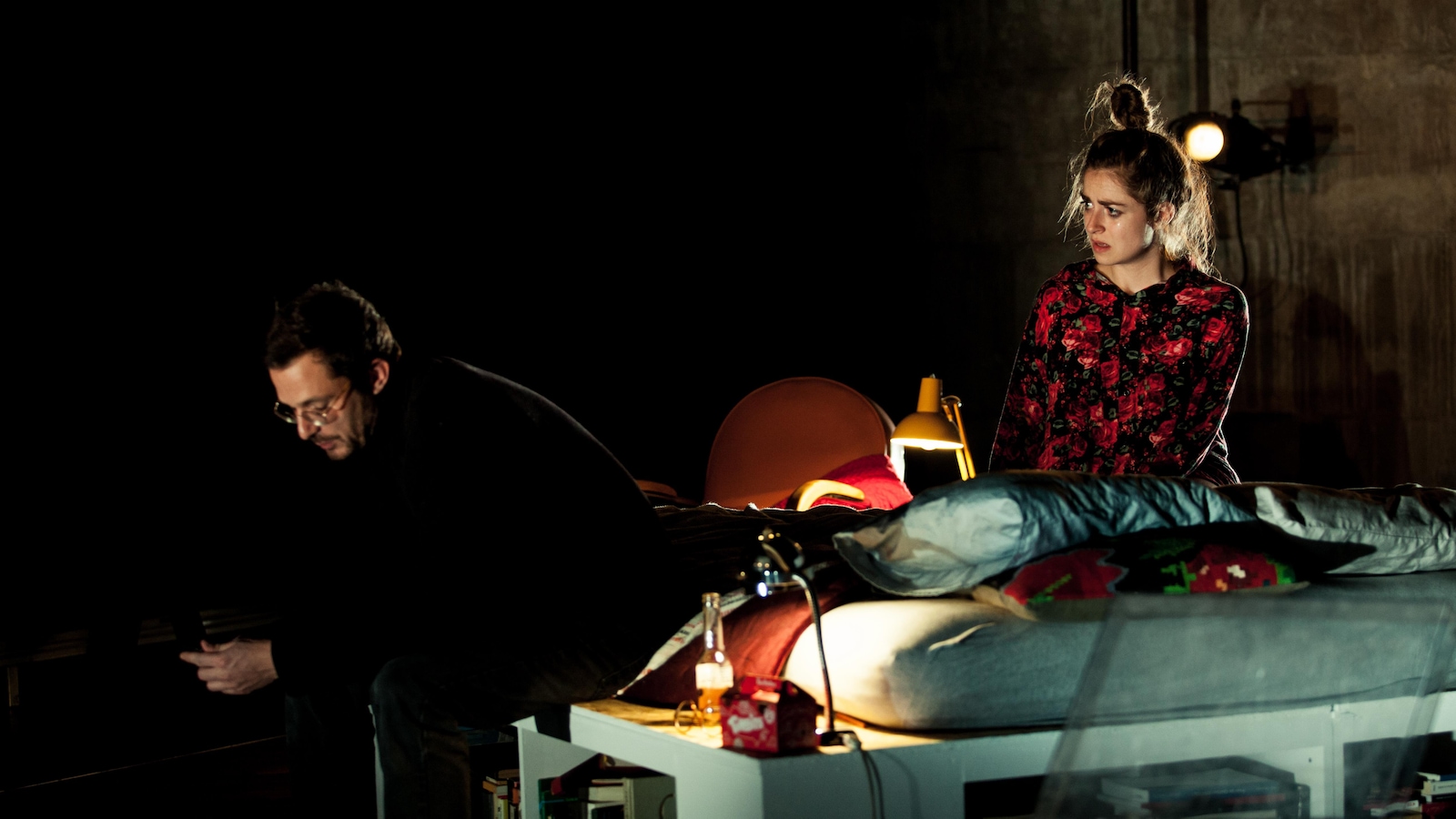 Un homme est assis au bout d'un lit, il regarde son téléphone dans ses mains, alors qu'une femme à genoux sur le lit le regarde. Un homme est assis au bout d'un lit, il regarde son téléphone dans ses mains, alors qu'une femme à genoux sur le lit le regarde.